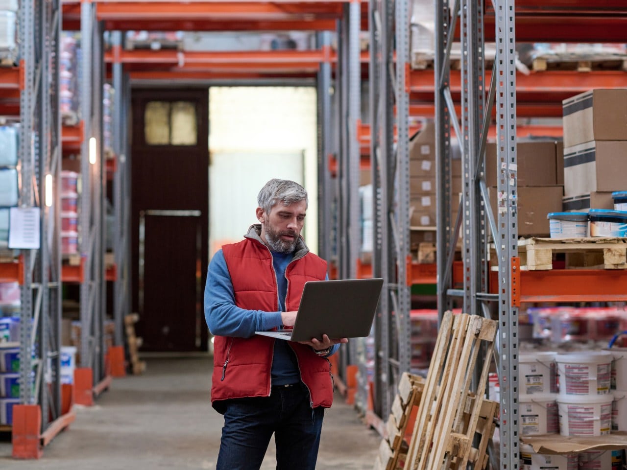 man in magazijn tussen stellingen werkt op laptop