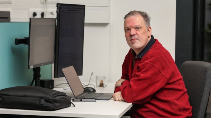 ICT medewerker zit bij bureau met meerdere schermen en kijkt in camera